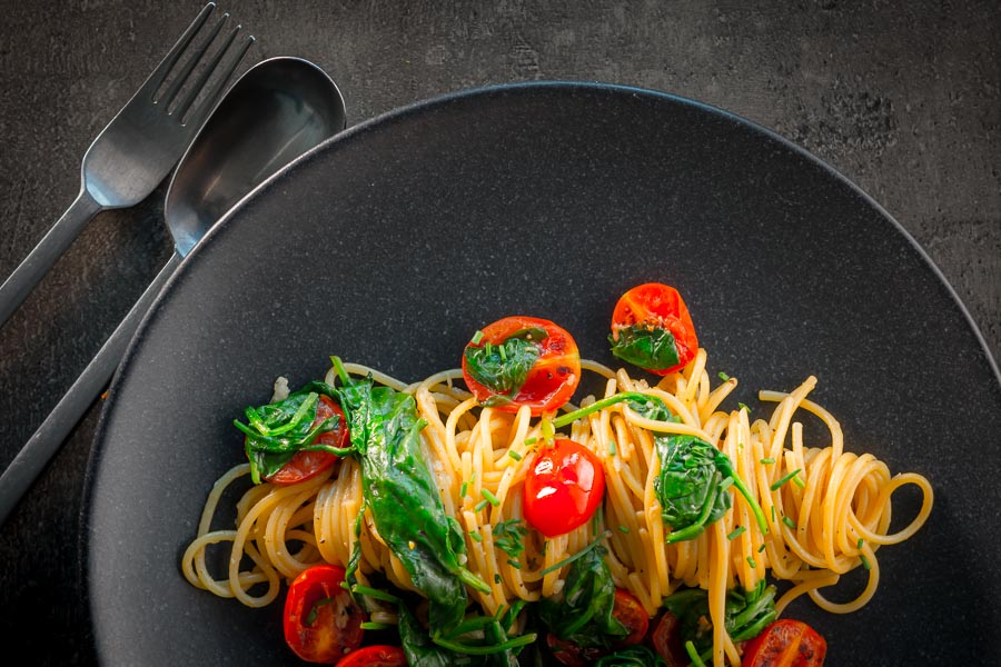 Landscape overhead image of Garlic pasta served in swirl on a dark plate with seared tomatoes and wilted spinach
