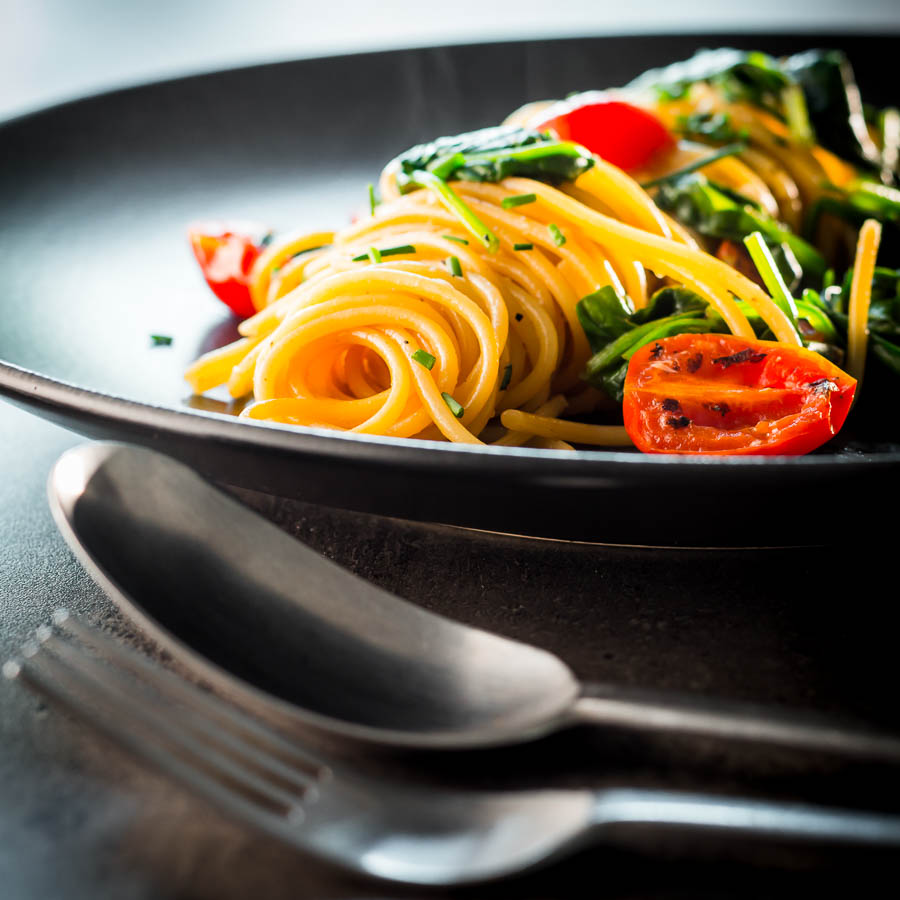 Square image of Garlic pasta served in swirl on a dark plate with seared tomatoes and wilted spinach