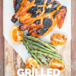 Portrait image of a Grilled Spatchcock Chicken served on a wooden chopping board with asparagus and grilled orange segments with Text