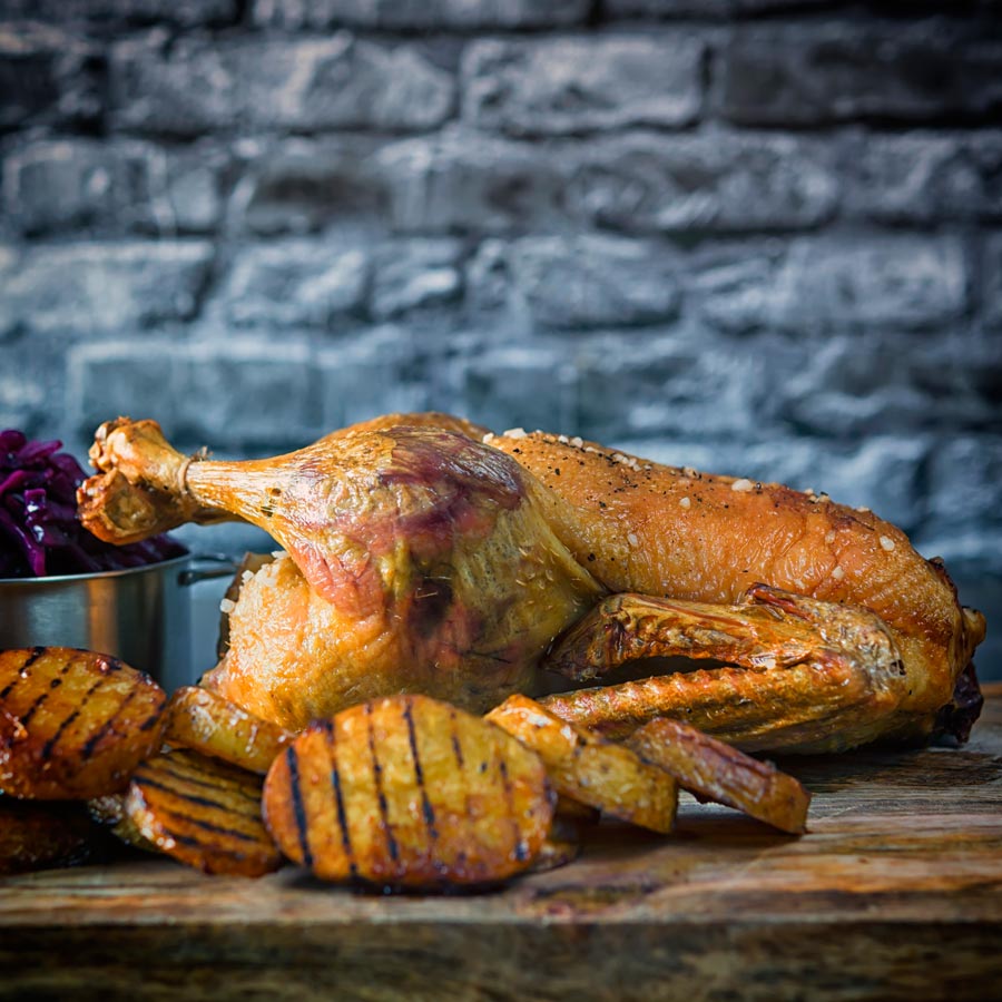 Square image of a whole roast duck with roasted scalloped potatoes served on a wooden serving board