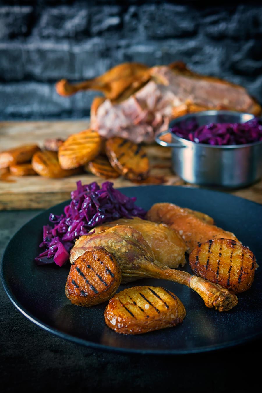 Portrait image of a duck leg from a whole roast duck served on a black plate with scalloped potatoes and red cabbage