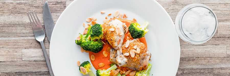 French trimmed chicken legs served on a romesco sauce with steamed broccoli and almonds, served on a white plate against a wood grain background.