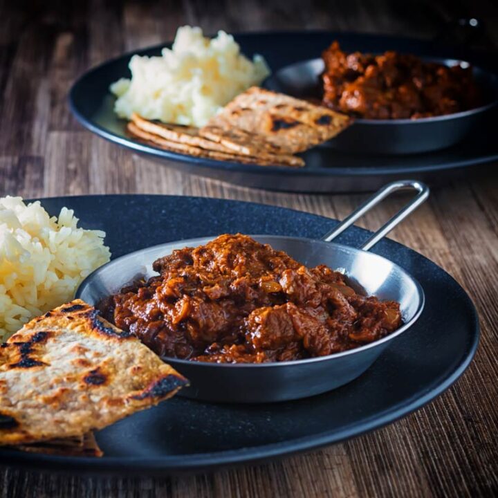 Square image of lamb madras curry served with white rice and chapatis served on a black plate