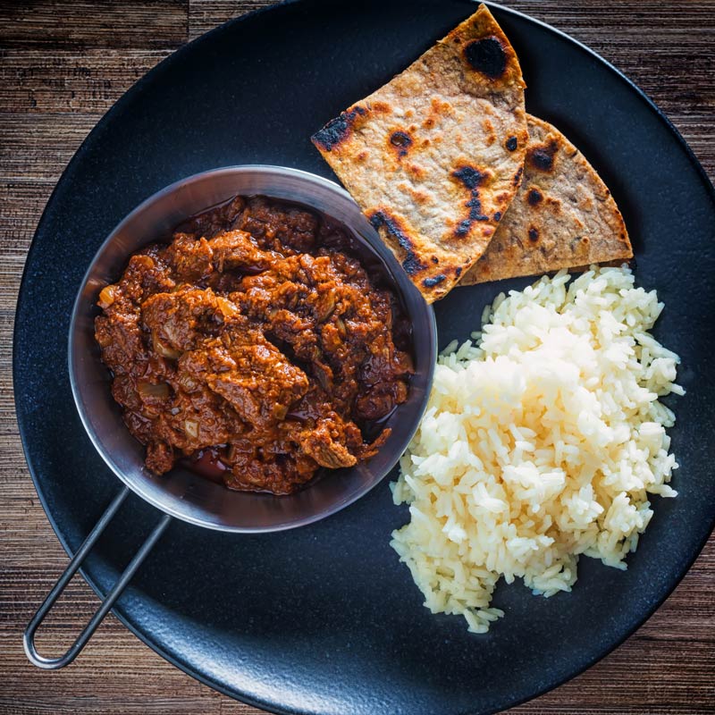 Square overhead image of lamb madras curry served with white rice and chapatis served on a black plate