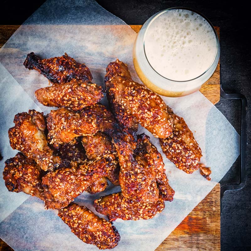 Square image taken from above of sticky spicy glazed buffalo chicken wings on a board with potato wedges and a beer