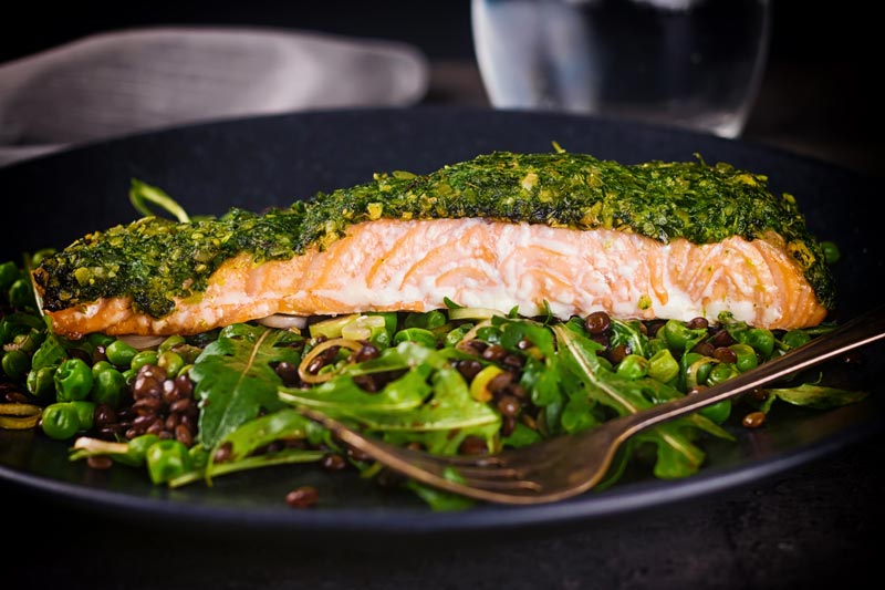Landscape image of a baked salmon filllet with a green herby crust with a fork showing the interior of the salmon