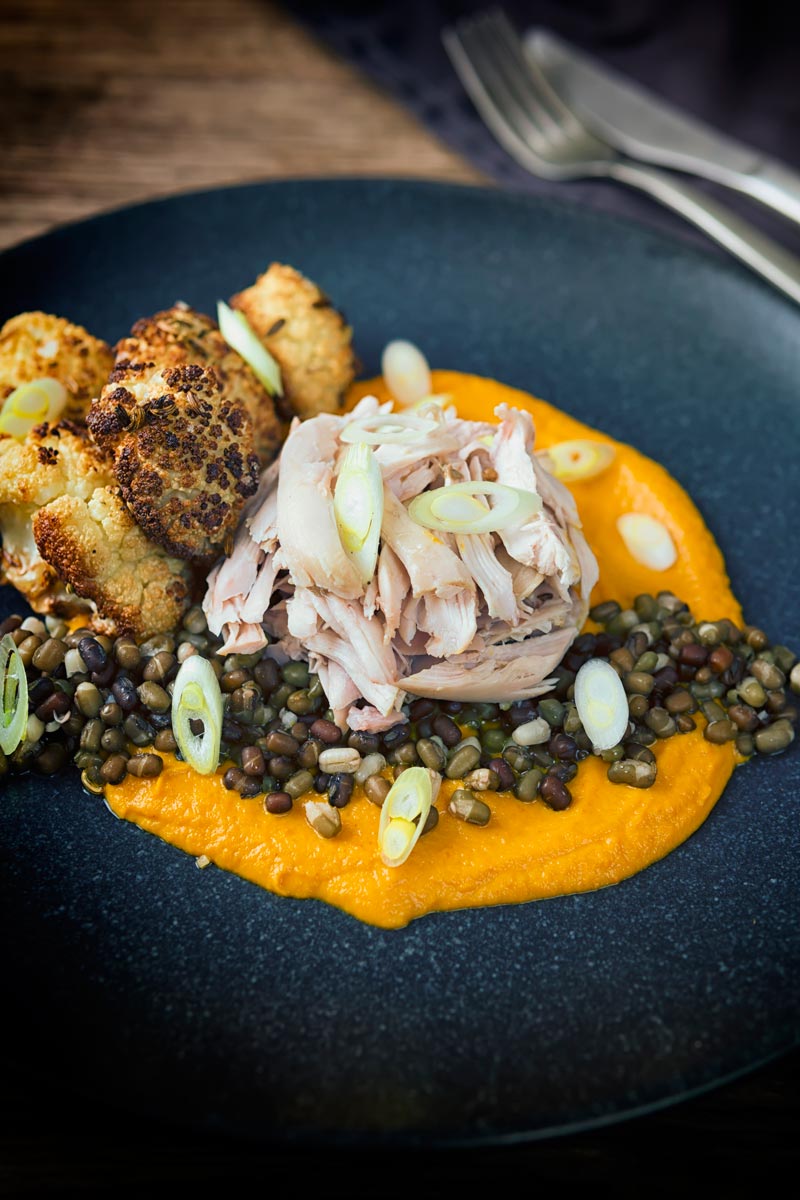 Portrait image of shredded slow cooked rabbit legs with mung beans, roasted cauliflower and spiced carrot puree on a dark plate