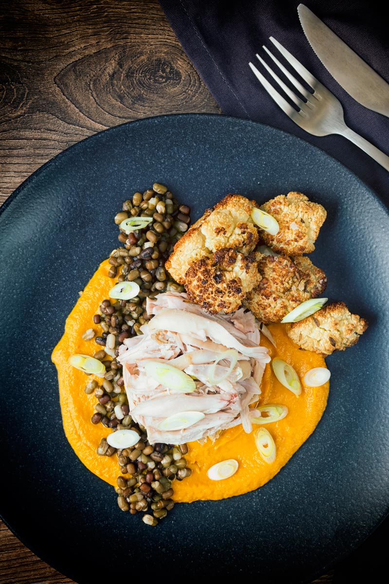 Portrait image taken from above of shredded slow cooked rabbit legs with mung beans, roasted cauliflower and spiced carrot puree on a dark plate