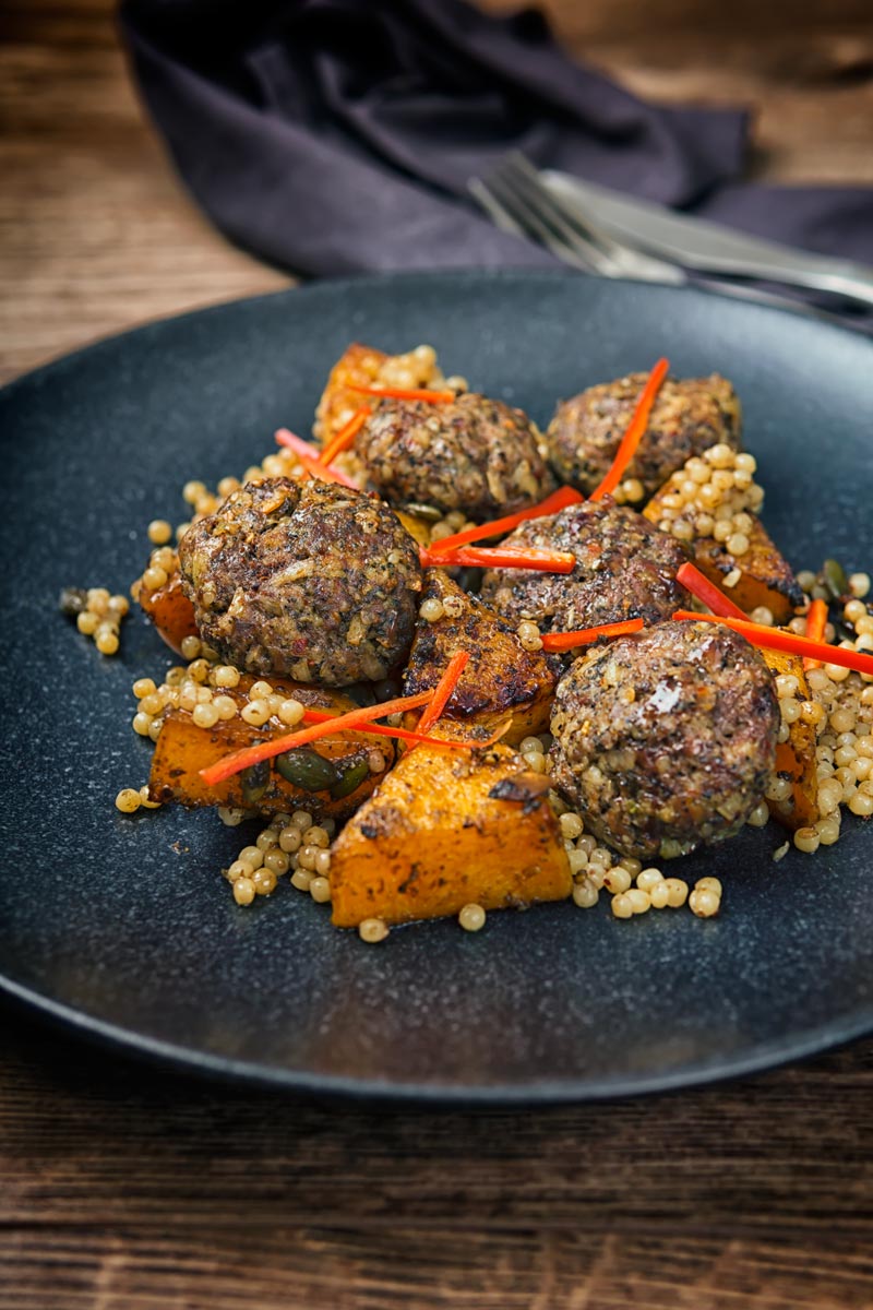 Portrait image of roasted butternut squash and Israeli couscous with Lamb kofta and chili batons served on a black plate