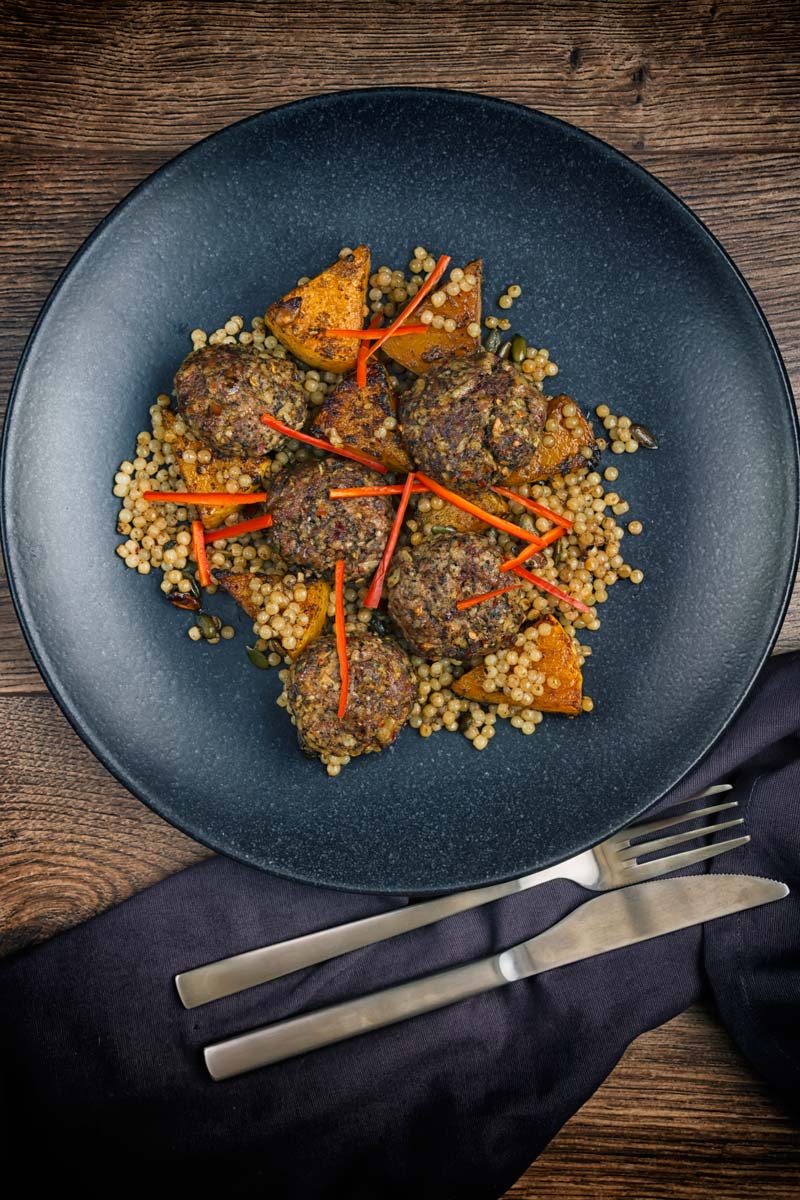 Portrait image of roasted butternut squash and Israeli couscous with Lamb kofta and chili batons served on a black plate taken from above