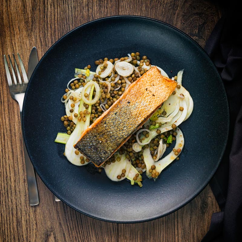 Square image of pan fried salmon with crispy skin served on lentils with pickled fennel on a black plate taken from above
