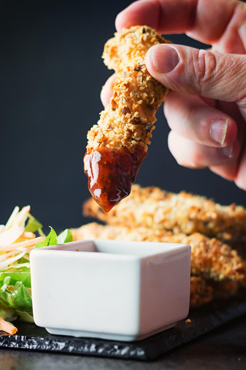 A baked crispy chicken tender dipped in a sauce against a dark backdrop
