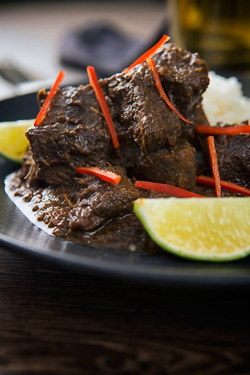 Close up portrait of an Indonesian Beef Rendang served on a black plate with rice, lime wedges and chilli ribbons.