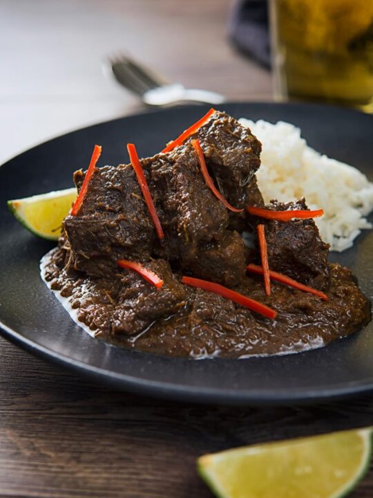 Portrait image of an Indonesian Beef Rendang served on a black plate with rice, lime wedges and chilli ribbons.
