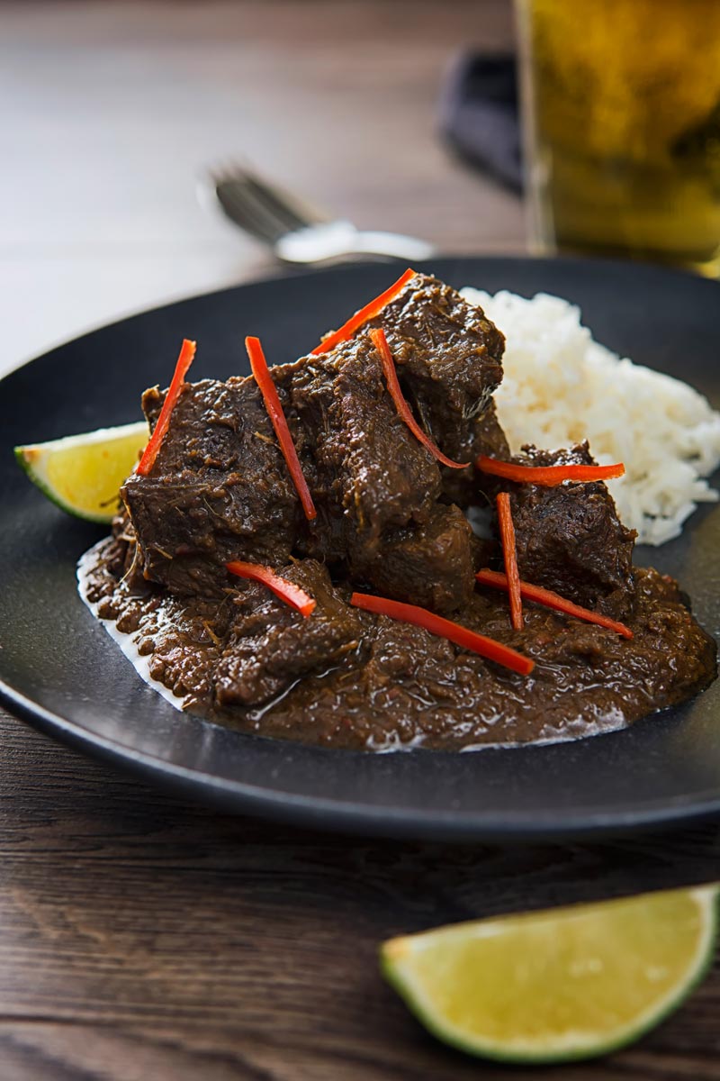 Portrait image of an Indonesian Beef Rendang served on a black plate with rice, lime wedges and chilli ribbons.