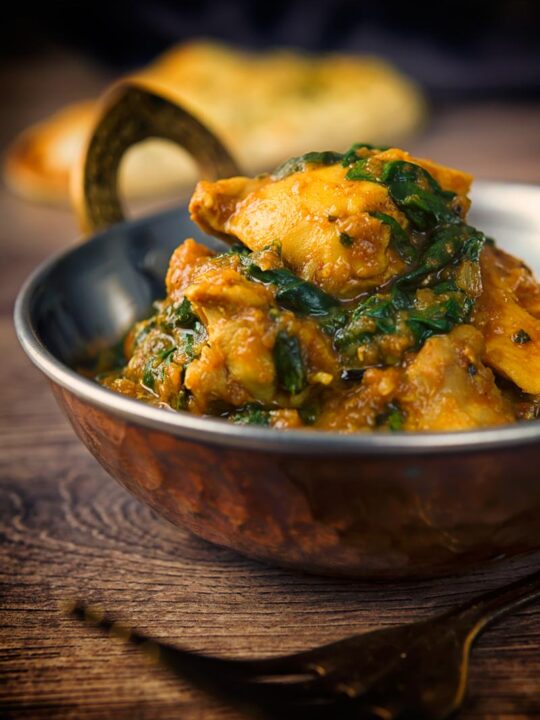 A tall image of a chicken saag curry in a 'brass' curry bowl on a wooden backdrop