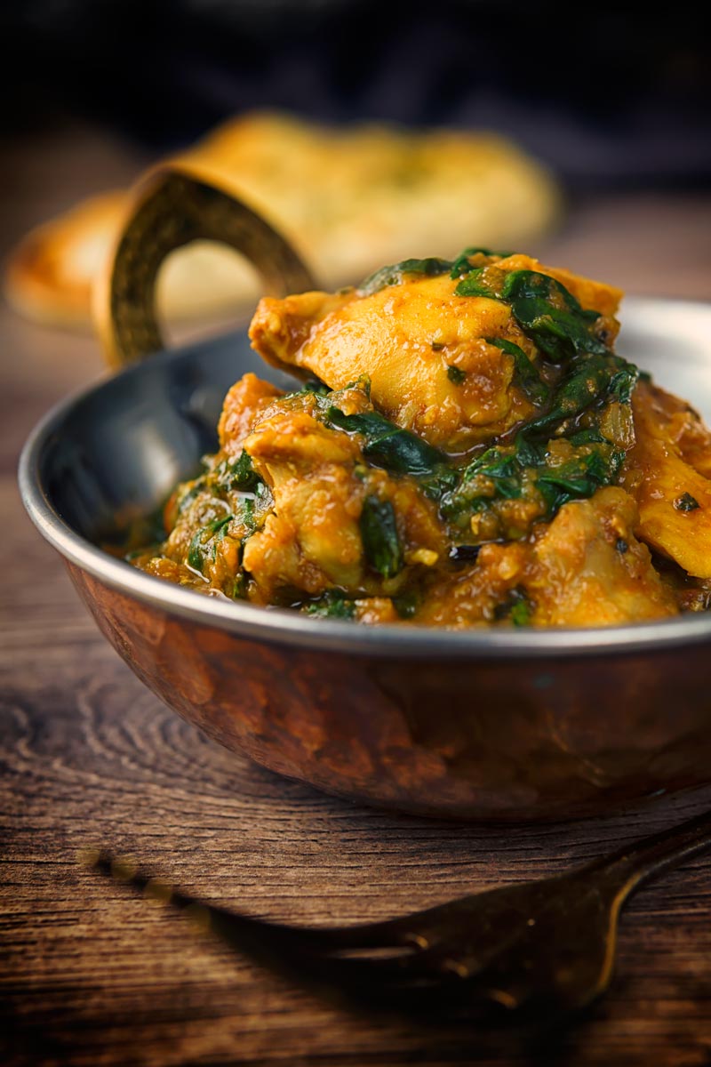 A tall image of a chicken saag curry in a 'brass' curry bowl on a wooden backdrop