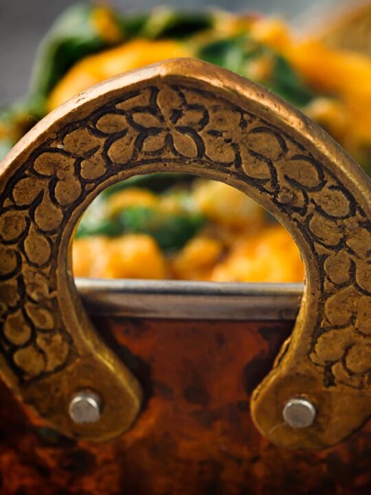 Close up image of the handle of a brass curry bowl with an out of focus curry in the background