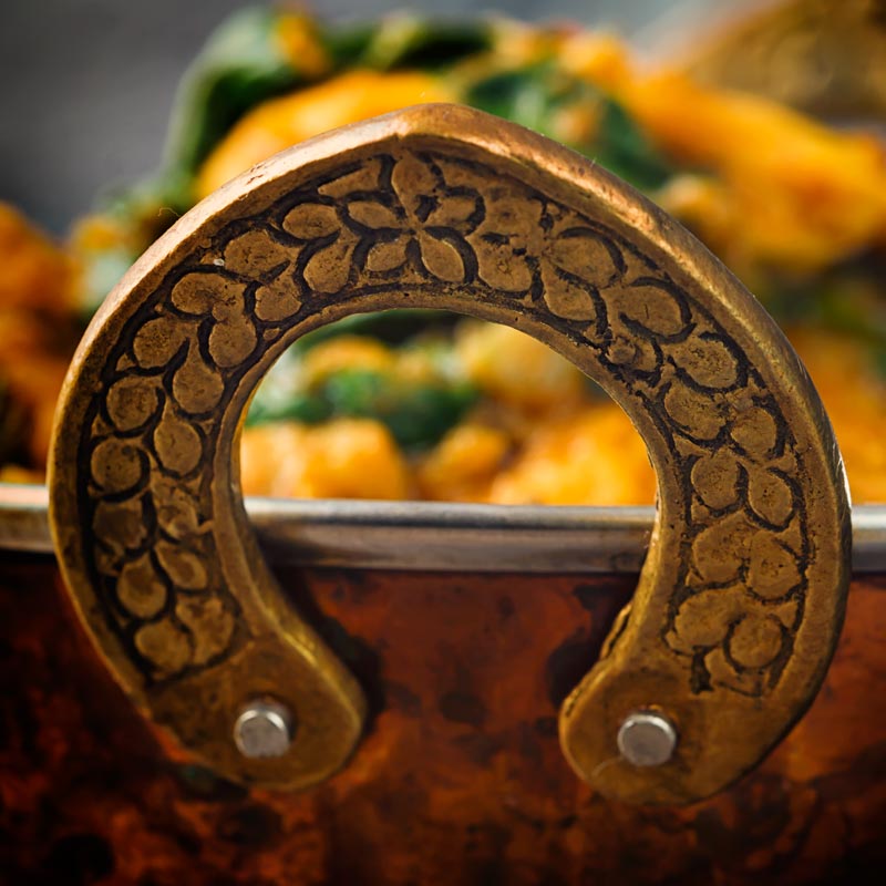 Close up image of the handle of a brass curry bowl with an out of focus curry in the background