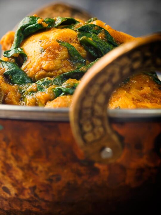 Close up image of chicken thighs and wilted spinach in a chicken saag curry in a brass curry bowl
