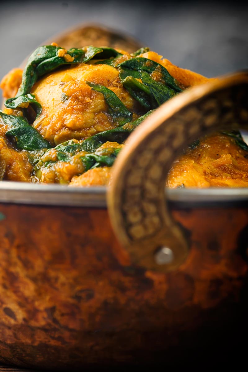 Close up image of chicken thighs and wilted spinach in a chicken saag curry in a brass curry bowl