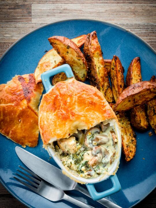 Tall image of a Chicken and mushroom pie on a blue plate with potato wedges with half of the pastry removed to expose the pie filling