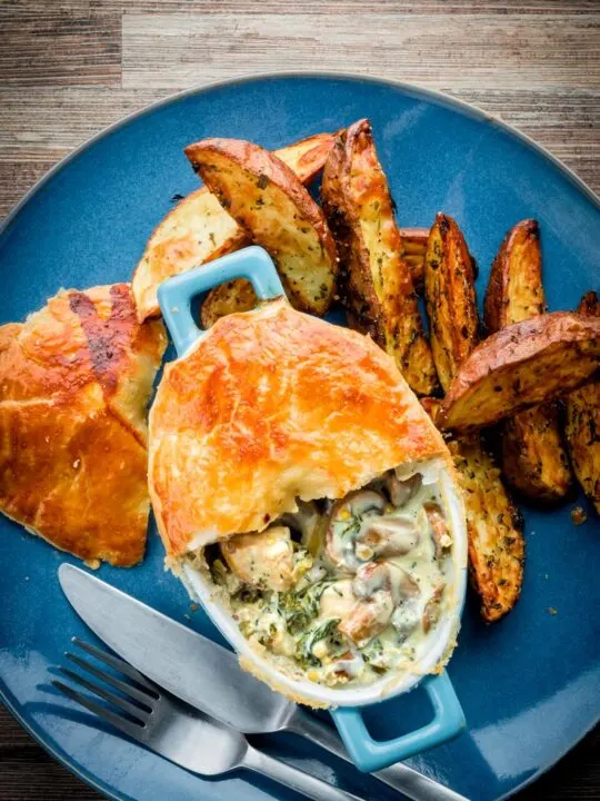 Tall image of a Chicken and mushroom pie on a blue plate with potato wedges with half of the pastry removed to expose the pie filling