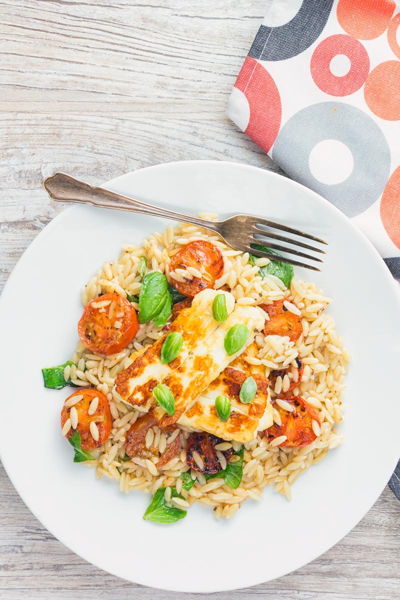 Tall image of seared Halloumi cheese sat on an orzo salad with tomatoes and basil taken from above.