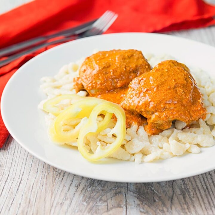 Square image of Hungarian Chicken Paprikash cooked with chicken thighs on buttered nokedli on a white plate
