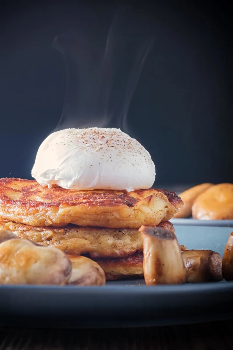 Stacked potato boxty with a steaming perfectly oval poached egg against a dark backdrop,