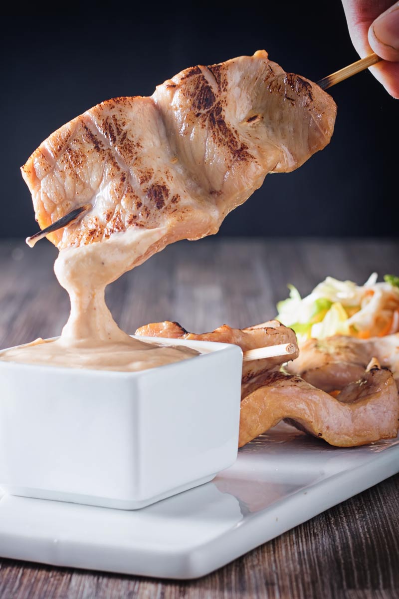 Tall image of pork satay being dipped into a peanut dipping sauce