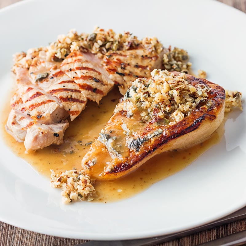 Square image of roasted pears with blue cheese and a savoury crumble and pork loin steak in a cider sauce on a white plate