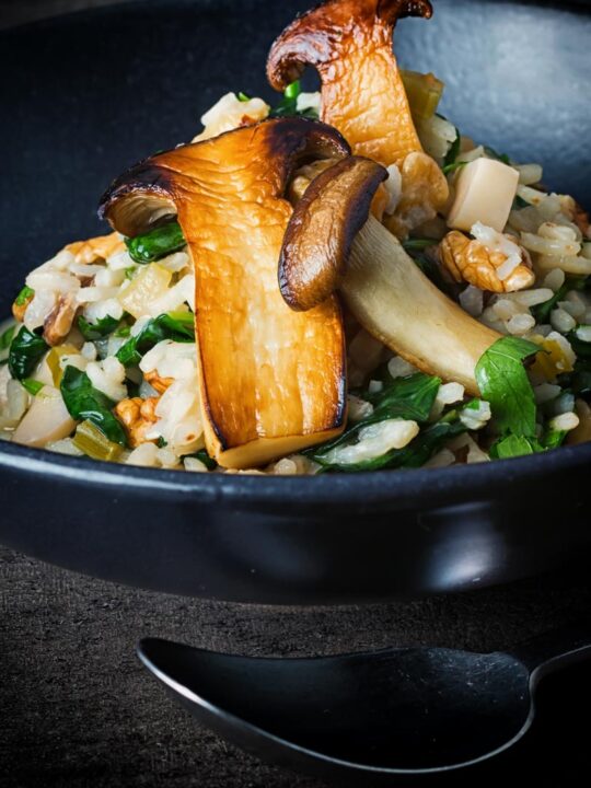Tall image of a spinach risotto with sauteed french horn or king oyster mushrooms in a black bowl