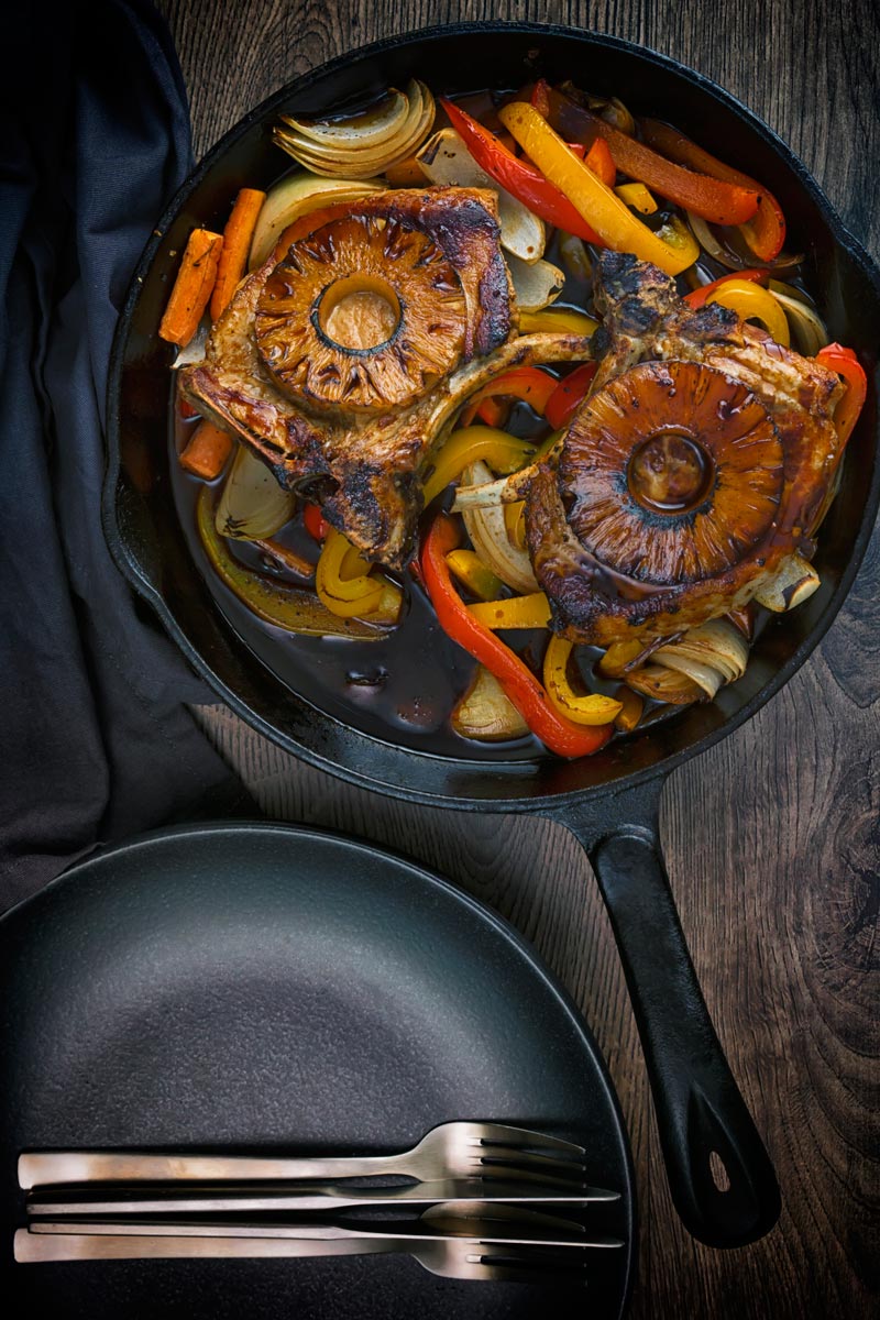 Portrait overhead image of sweet and sour pork chops cooked and served in a cast iron skillet with vegetables and a pineapple ring