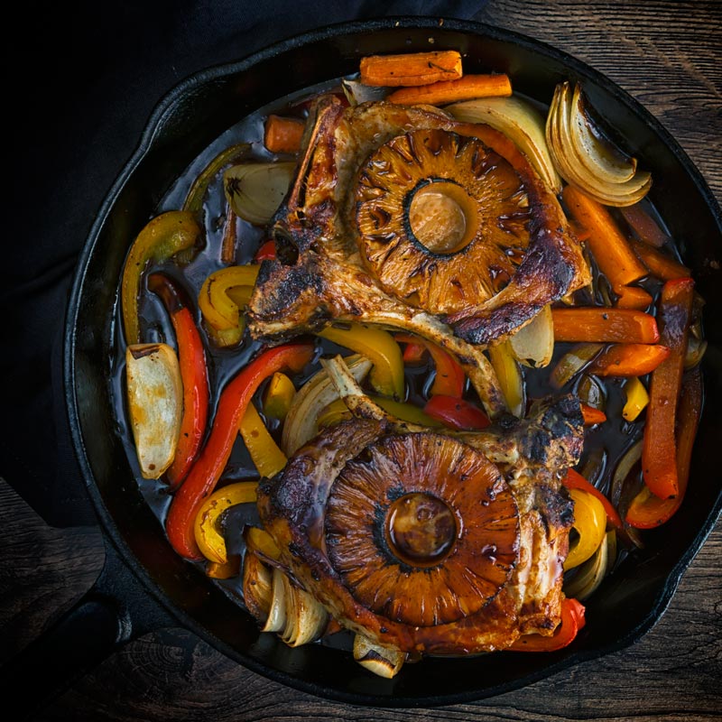 Two sweet and sour pork chops topped with pineapple rings on a bed of sweet peppers and onions in a cast iron pan