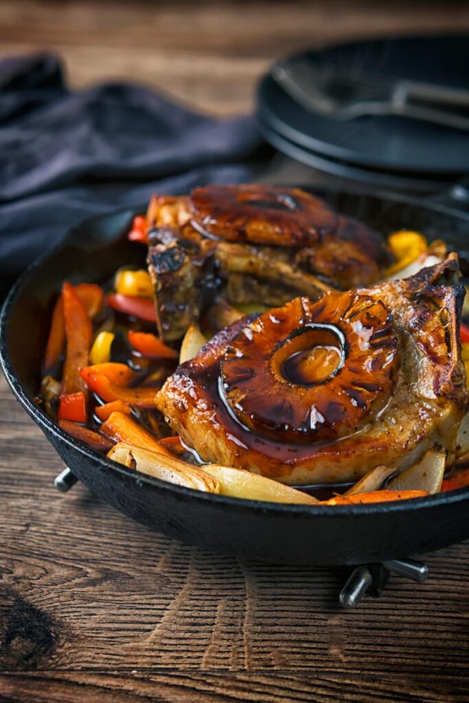 Portrait image of sweet and sour pork chops cooked and served in a cast iron skillet on a rustic wooden table with vegetables and a pineapple ring