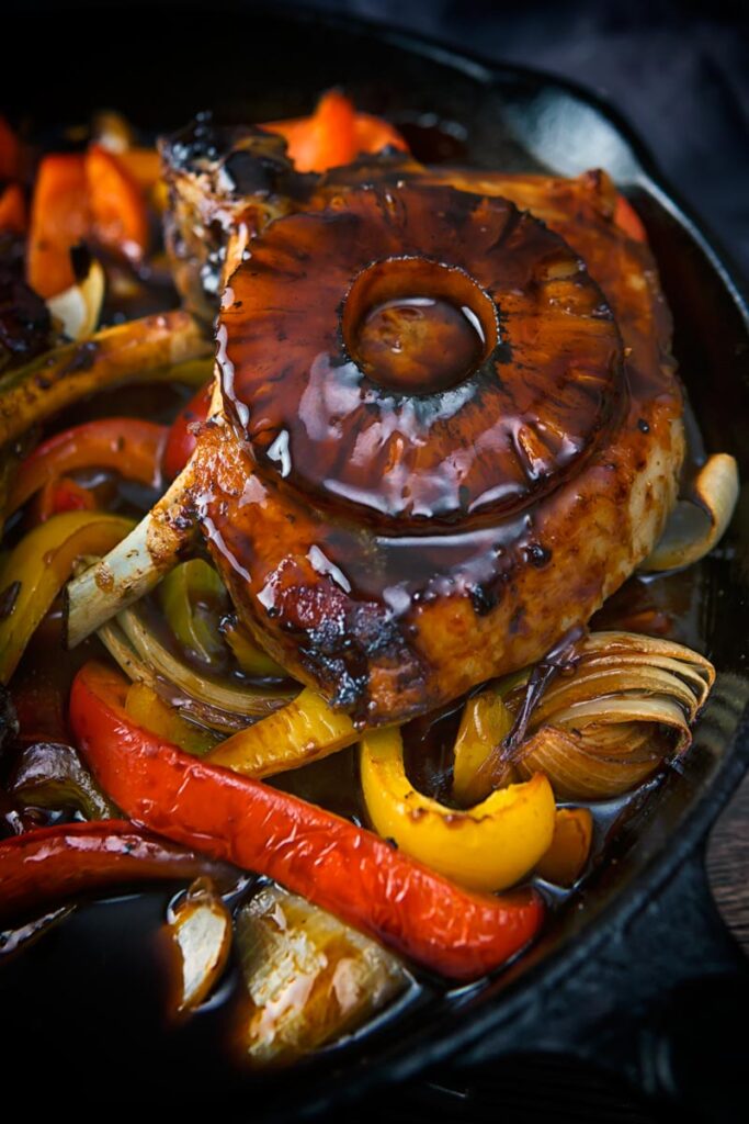 Portrait image of sweet and sour pork chops cooked and served in a cast iron skillet with vegetables and a pineapple ring