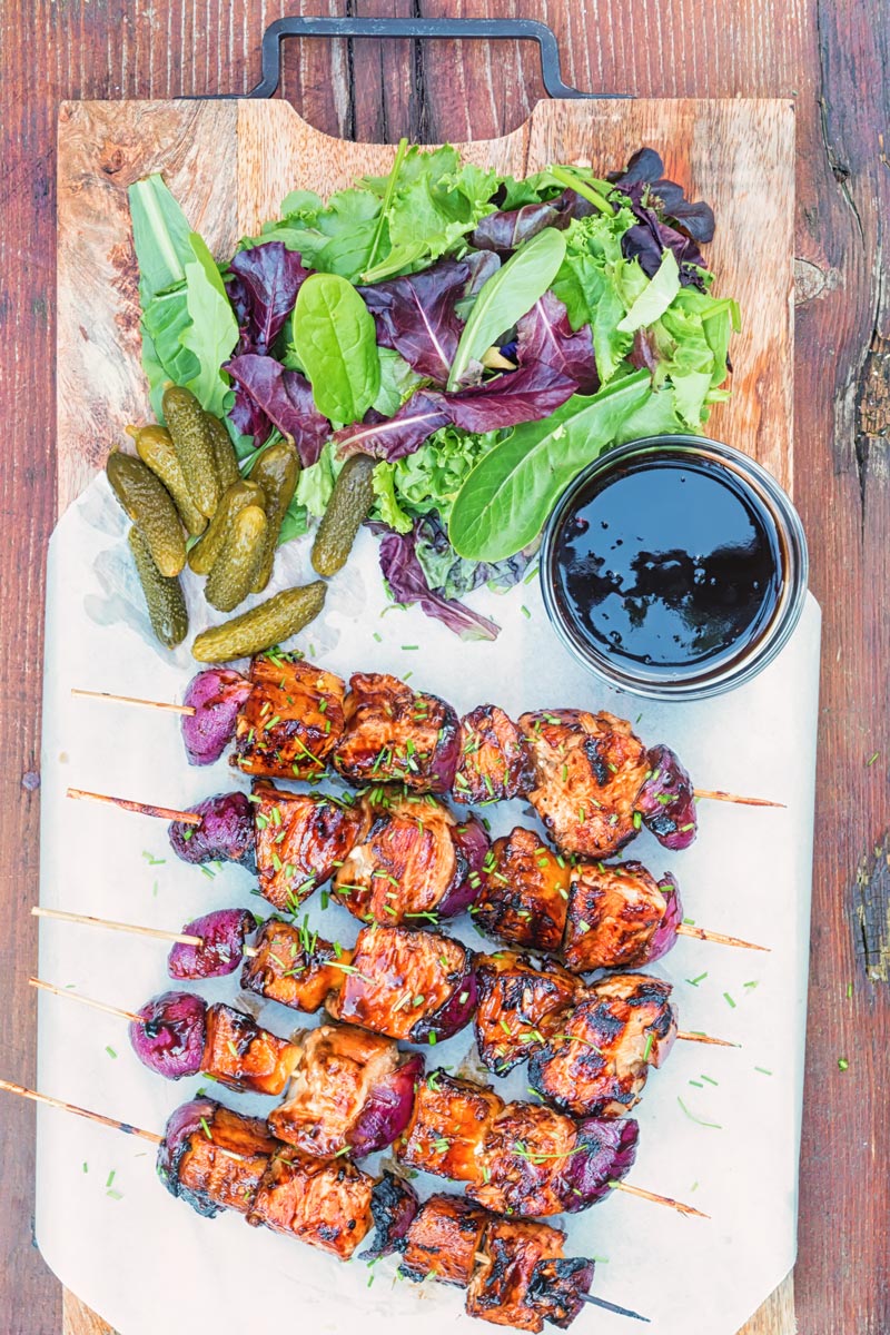 Overhead portrait image of bbq pineapple chicken kebabs served on a chopping board with a BBQ sauce, side salad and pickles