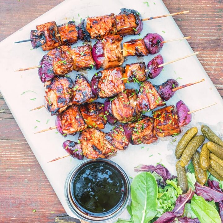 Square portrait image of bbq pineapple chicken kebabs served on a chopping board with a BBQ sauce, side salad and pickles