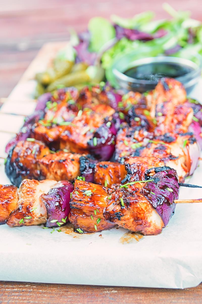 Portrait image of bbq pineapple chicken kebabs served on a chopping board with a BBQ sauce, side salad and pickles