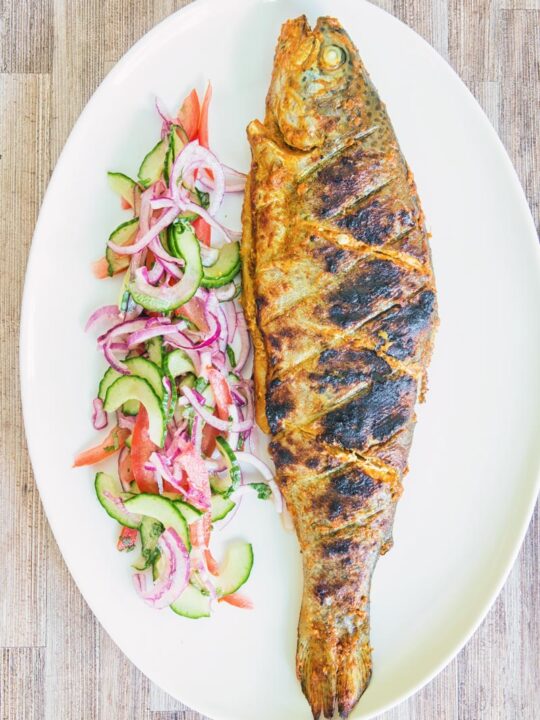 A whole BBQ tandoori fish with a kachumber salad on an oval plate taken from above