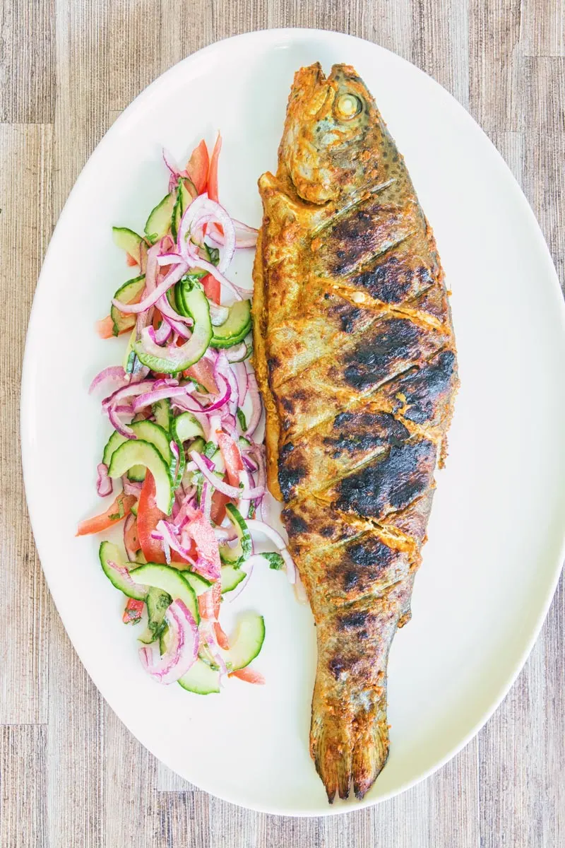 A whole BBQ tandoori fish with a kachumber salad on an oval plate taken from above