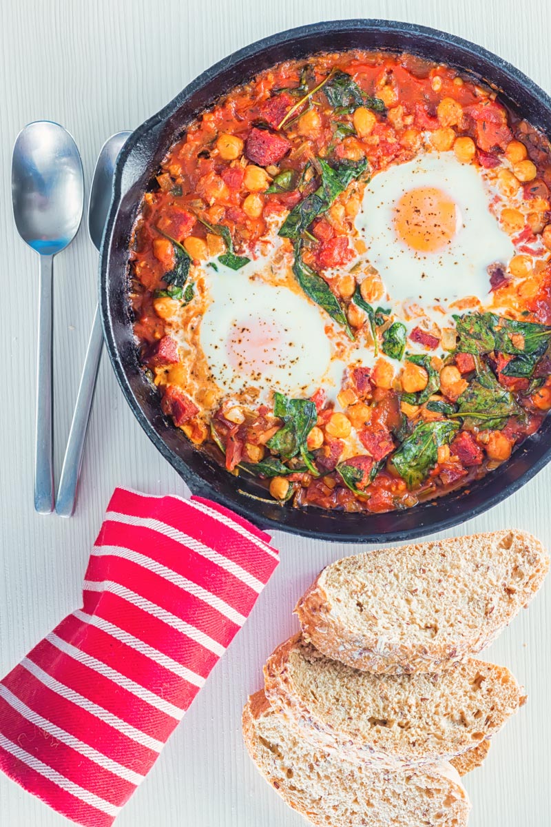 Overhead portrait image of baked chickpeas with tomato and eggs served and cooked in a cast iron skillet served with seeded bread