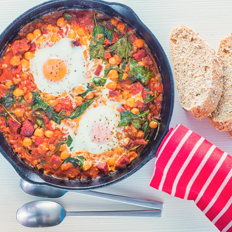 Overhead square image of baked chickpeas with tomato and eggs served and cooked in a cast iron skillet served with seeded bread