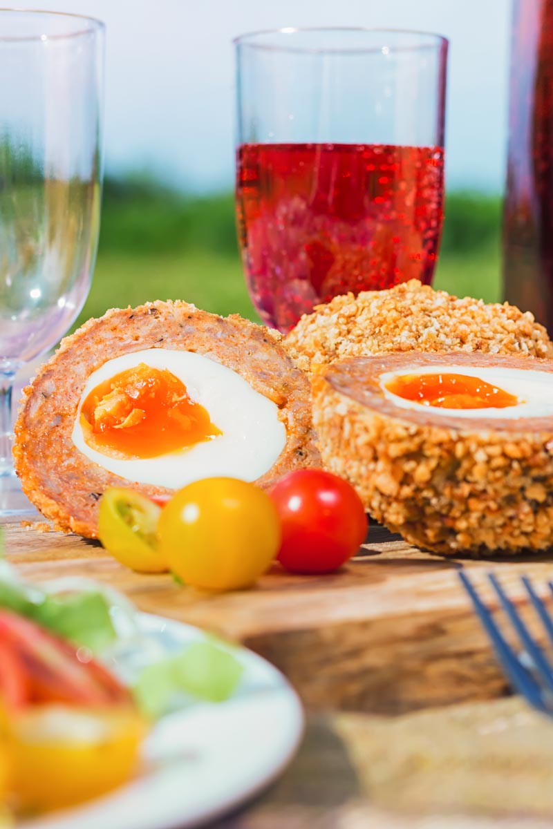 Tall close up image of a baked scotch egg with a perfect jammy yolk in a picnic setting