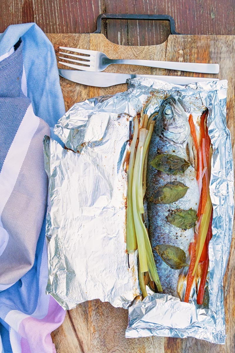 Portrait image of baked fish in foil with vegetable ribbons and basil leaves on a wooden chopping board