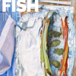 Portrait image of baked fish in foil with vegetable ribbons and basil leaves on a wooden chopping board with text