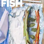 Portrait image of baked fish in foil with vegetable ribbons and basil leaves on a wooden chopping board with text