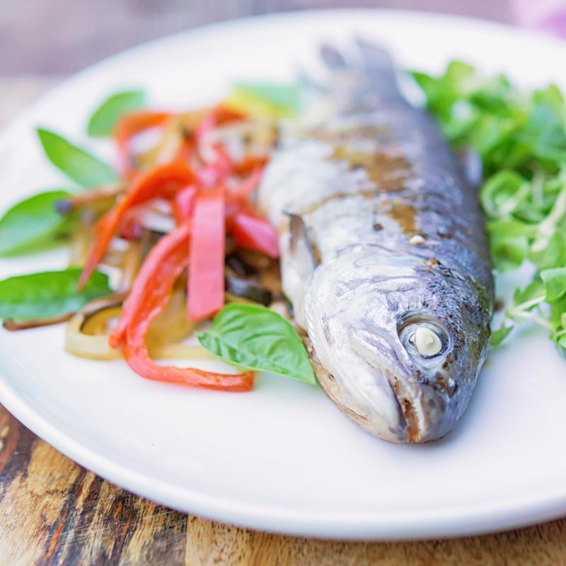 Square image of a whole cooked trout baked in foil with vegetable ribbons and side salad on a white plate
