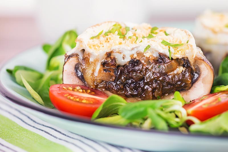 Landscape image of stuffed portobello mushrooms with goats cheese and a breadcrumb topping cut open to show a caramelised onion centre served with a green salad and tomatoes